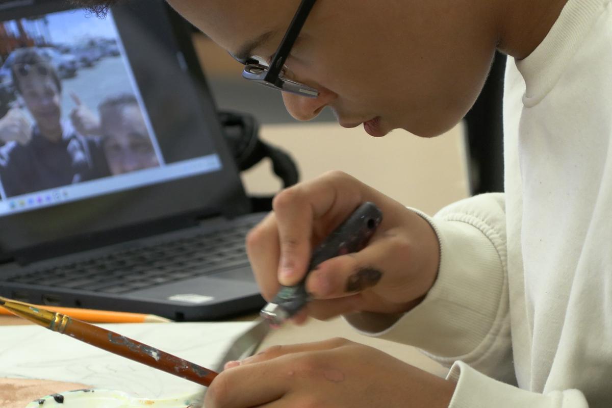 High schooler Xavier King leans close to a portrait he's painting next to his laptop.